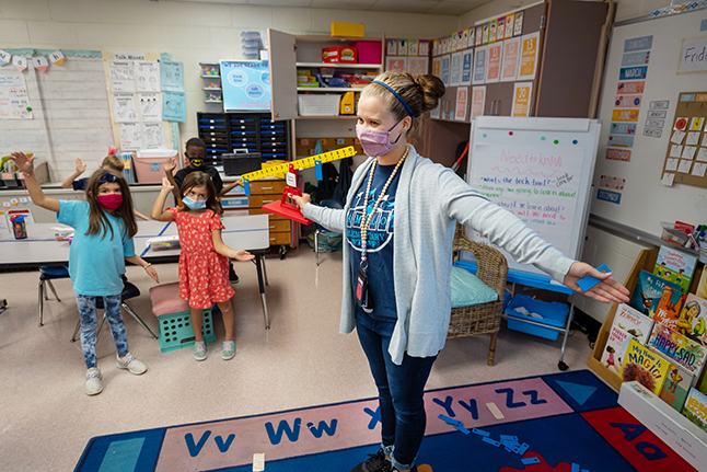A teacher with arms outstretched shows her students a math problem on a scale. 