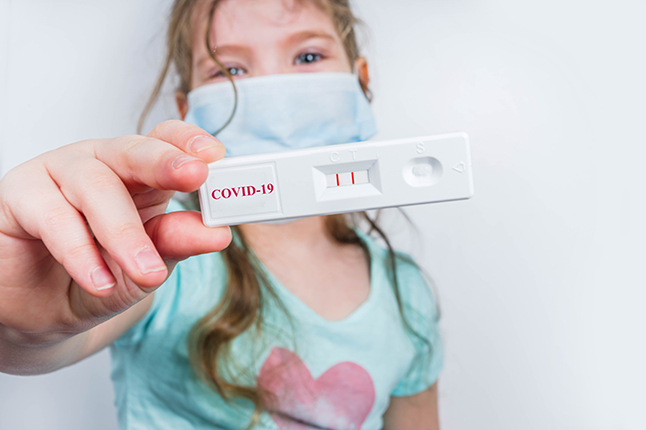 A young girl wearing a mask holds up a COVID test