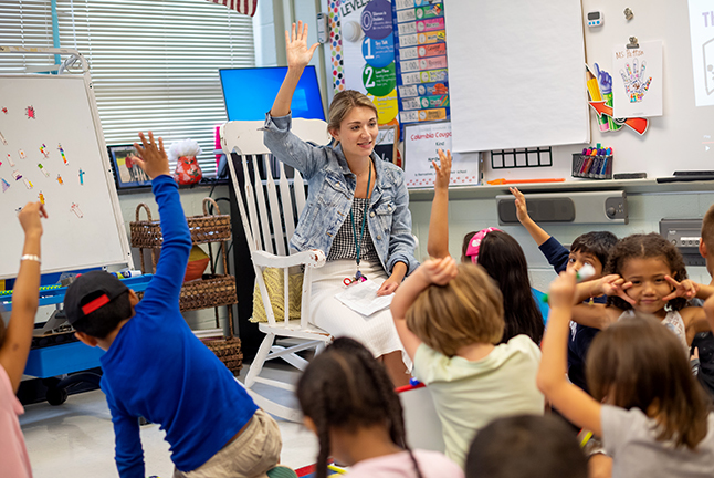 A teacher in a rocking chair raises her hand before young children with their hands raised. 