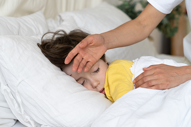 A student lies in bed while an adult feels their forehead with the back of their hand. 