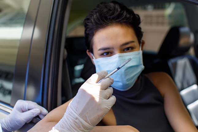 A masked person sitting in a car looks at a gloved hand holding an injection