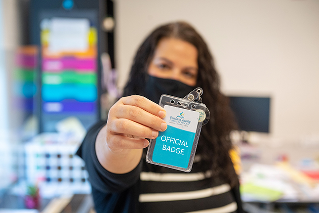 A masked adult holds up a work ID badge