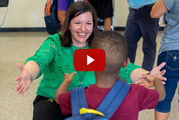 A teacher holds her arms out greeting a young boy, with YouTube play button overlay