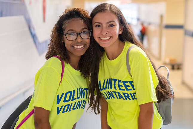 Two proud and smiling women wearing bright yellow Volunteer/Voluntario shirts.