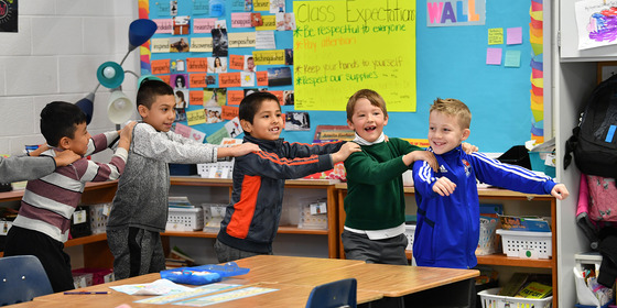 Elementary age students in connected front to back line with hands their leaders shoulders as walk around a classroom.
