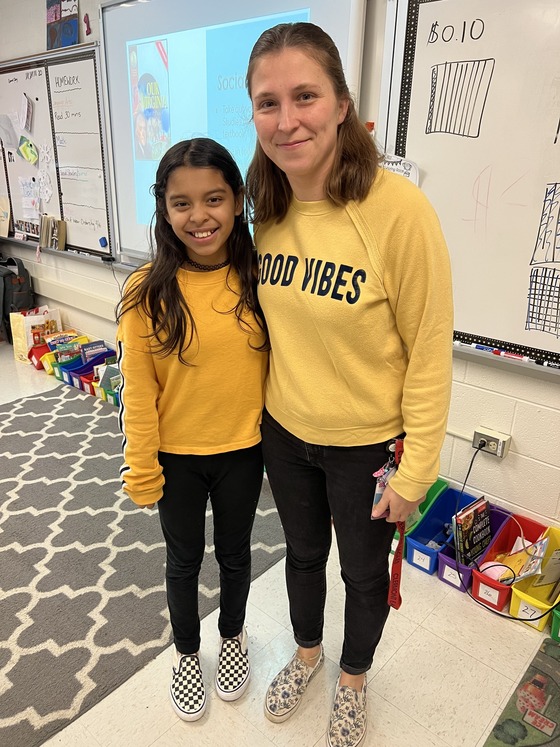 A student and staff member, both dressed in black pants, yellow shirts, and black and white canvas shoes, pose together. 