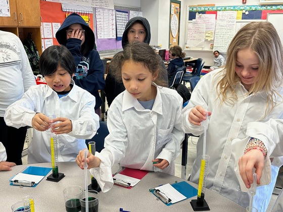 students in white lab coats conduct a science experiment using pipettes and beakers
