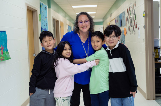 Teacher smiles while four students are hugging her. 