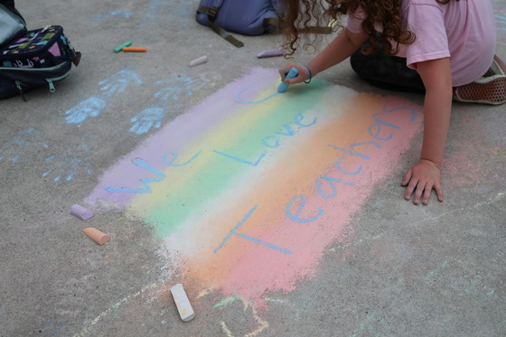 A childs hands writing the words "We Love Teachers" on a rainbow background in sidewalk chalk.