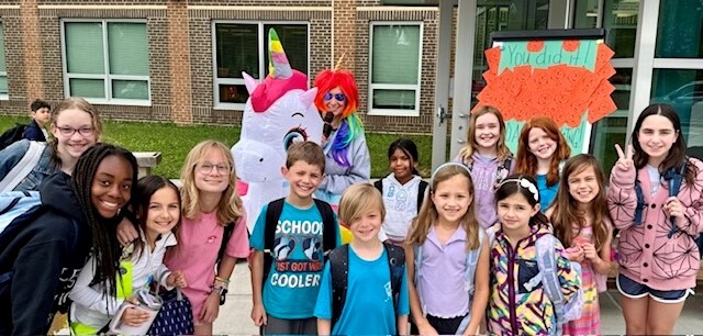 Principal Salata, wearing a rainbow wig and inflatable unicorn costume poses with students holding a microphone. 
