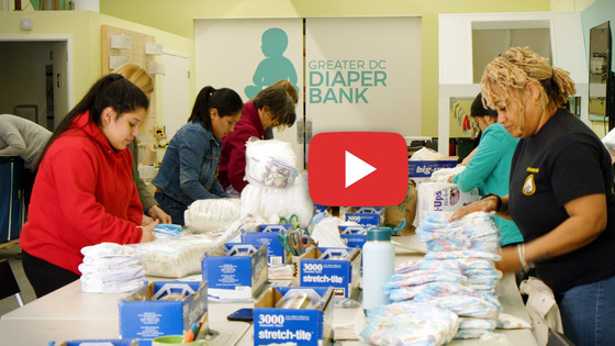 Greater DC Diaper Bank as volunteers pack and prepare diapers. Image includes a red and white video play button.
