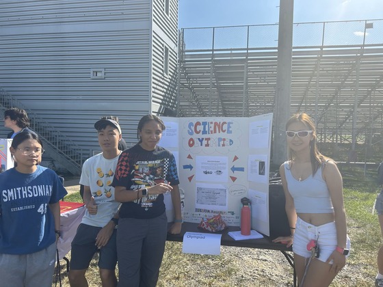 Students standing, outside, on either side of a posterboard that says science olympiad