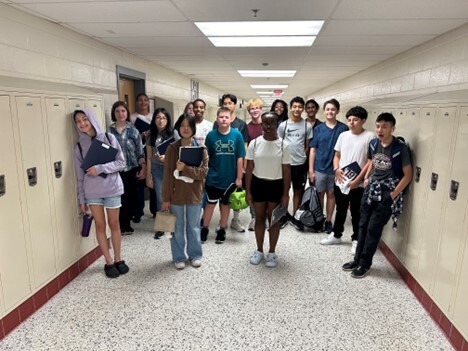 17 students standing in the hallway holding books and smiling