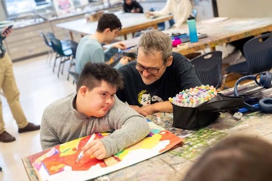 teacher smiles next to student who is creating a colorful drawing