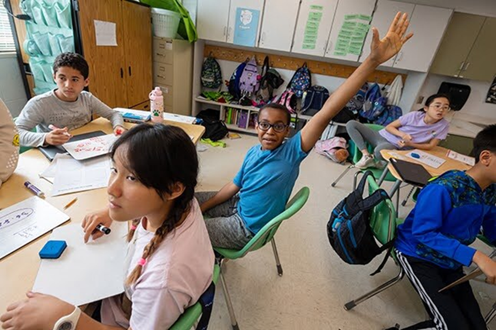 Students sitting around tables in classroom with one student's hand raised