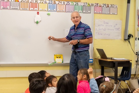 teacher writes on a whiteboard while students sit on the carpet in front of him