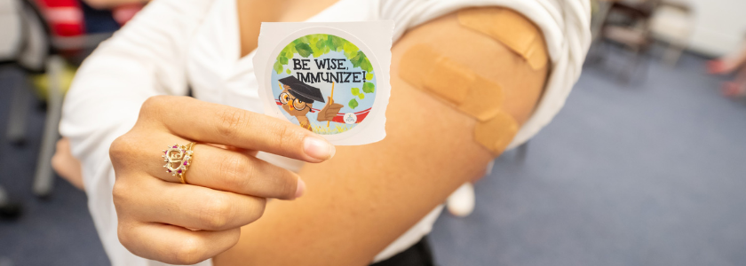 photo of arm with bandaid and sticker that reads "Be wise, Immunize"