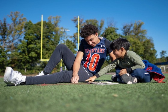 high school student reading to an elementary school students while sitting on the football field