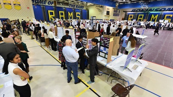 Students standing at tables presenting their findings to judges in gymnasium