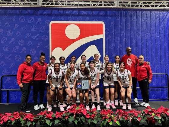 Girls basketball team standing together on a stage with coaches in front of a red, blue, and white sign