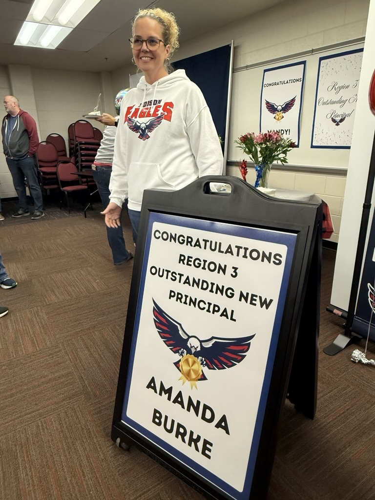 Principal standing behind a sign that says congratulations region 3 outstanding new principal Amanda Burke