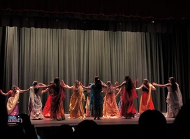 A group of dancers in colorful traditional saris perform on stage, facing a green curtain backdrop.
