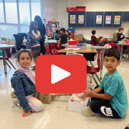 Kindergarten classroom with children engaged in activities. A girl and boy sit on the floor smiling. A play button icon indicates a video thumbnail.