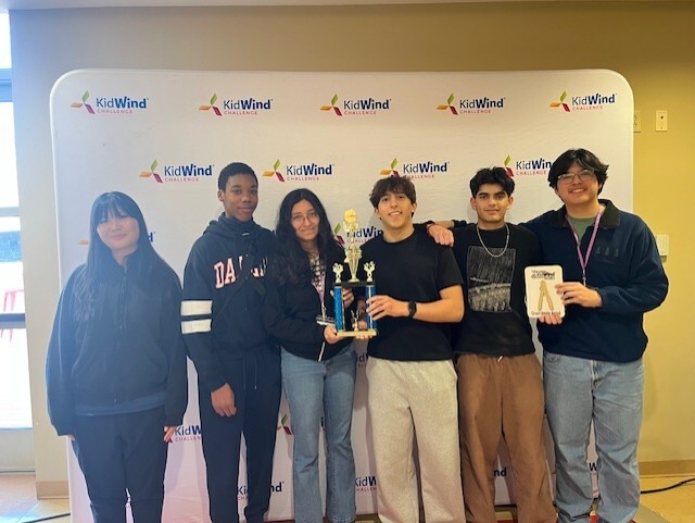 A group of six young people standing in front of a KidWind logo backdrop, some holding trophies.
