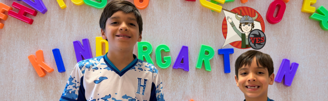 Two students smiling beneath colorful letters spelling 'KINDERGARTEN' on wall, wearing blue and white patterned shirts