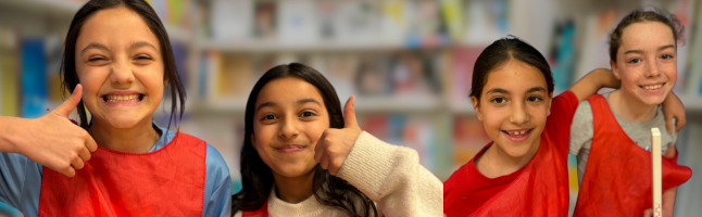 Four students giving thumbs up and smiling, wearing red and beige tops, with bookshelves visible in background