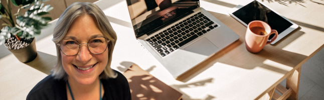Woman wearing glasses smiling at desk workspace with laptop, orange coffee mug, and potted plant against white surface with natural lighting