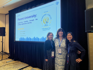 Three women standing together, smiling, in front of a presentation screen at a conference.