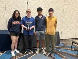 Four young individuals stand in a row; one holds a trophy. They are dressed in casual attire, with a patterned wall in the background.