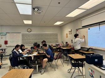 One student writing on a white board, while a group of four students sit at a desk