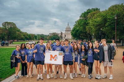 Group of young adults posing on a path with the U.S. Capitol in the background, holding a sign with "MC" visible.
