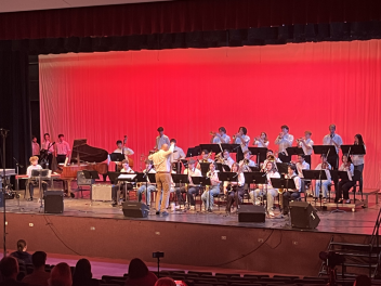 Large musical ensemble performing on stage with red background, led by a conductor.