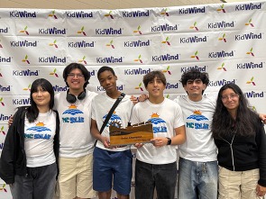 A group of six people in matching T-shirts holding a "Solar Champion" trophy in front of a "KidWind" logo backdrop.