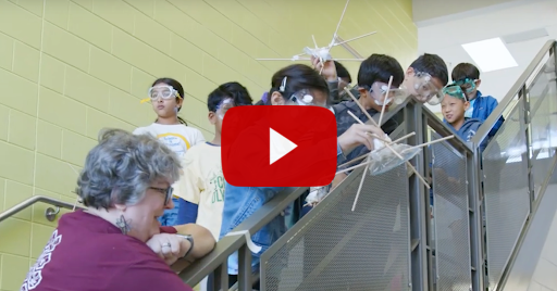 Screenshot: Students and a teacher holding objects over a railing preparing to drop them in a science experiment
