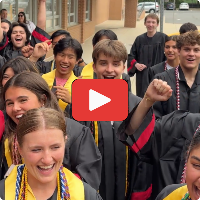 Group of graduates in black gowns and colorful honor cords celebrating outside building. Red play button overlay in center