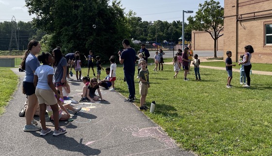 5th graders and kindergarten students playing outside together with bubbles and sidewalk chalk