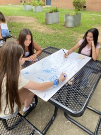 Three young women writing on a whiteboard at a picnic table outdoors.