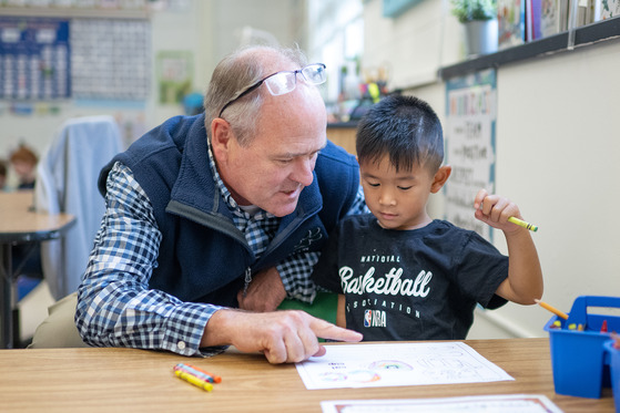 A man points to a worksheet a young student is working on with a crayon