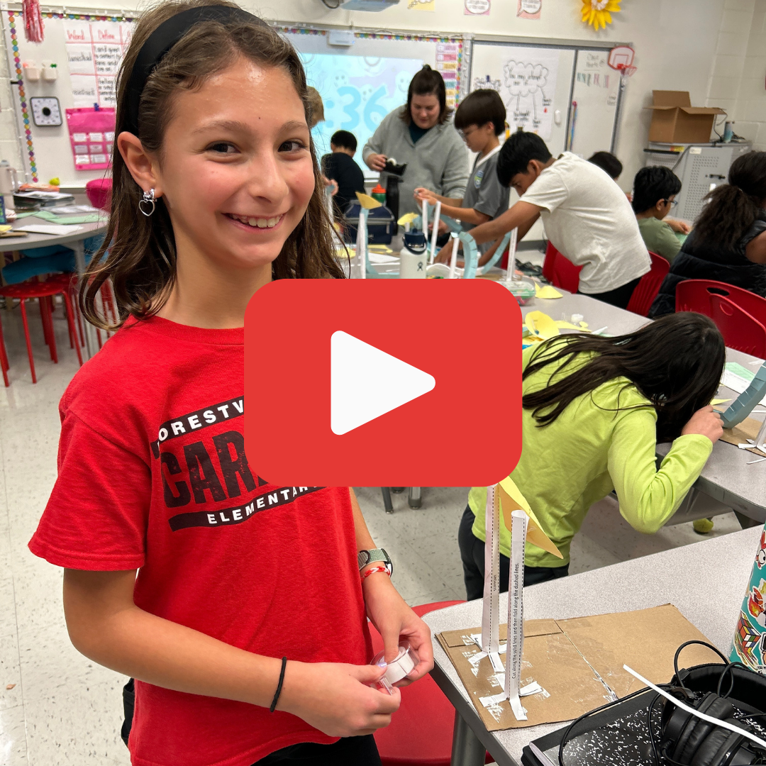 A young girl in a Forestville T-Shirt standing next to a paper roller coaster, in a classroom. A red play button is over the photo