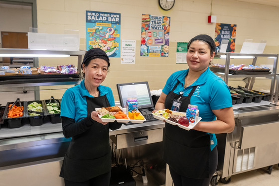 Two cafeteria staff hold trays of food