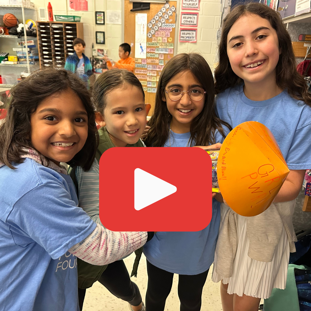 Four smiling students stand together in a classroom, holding an orange paper project shaped like a cone.