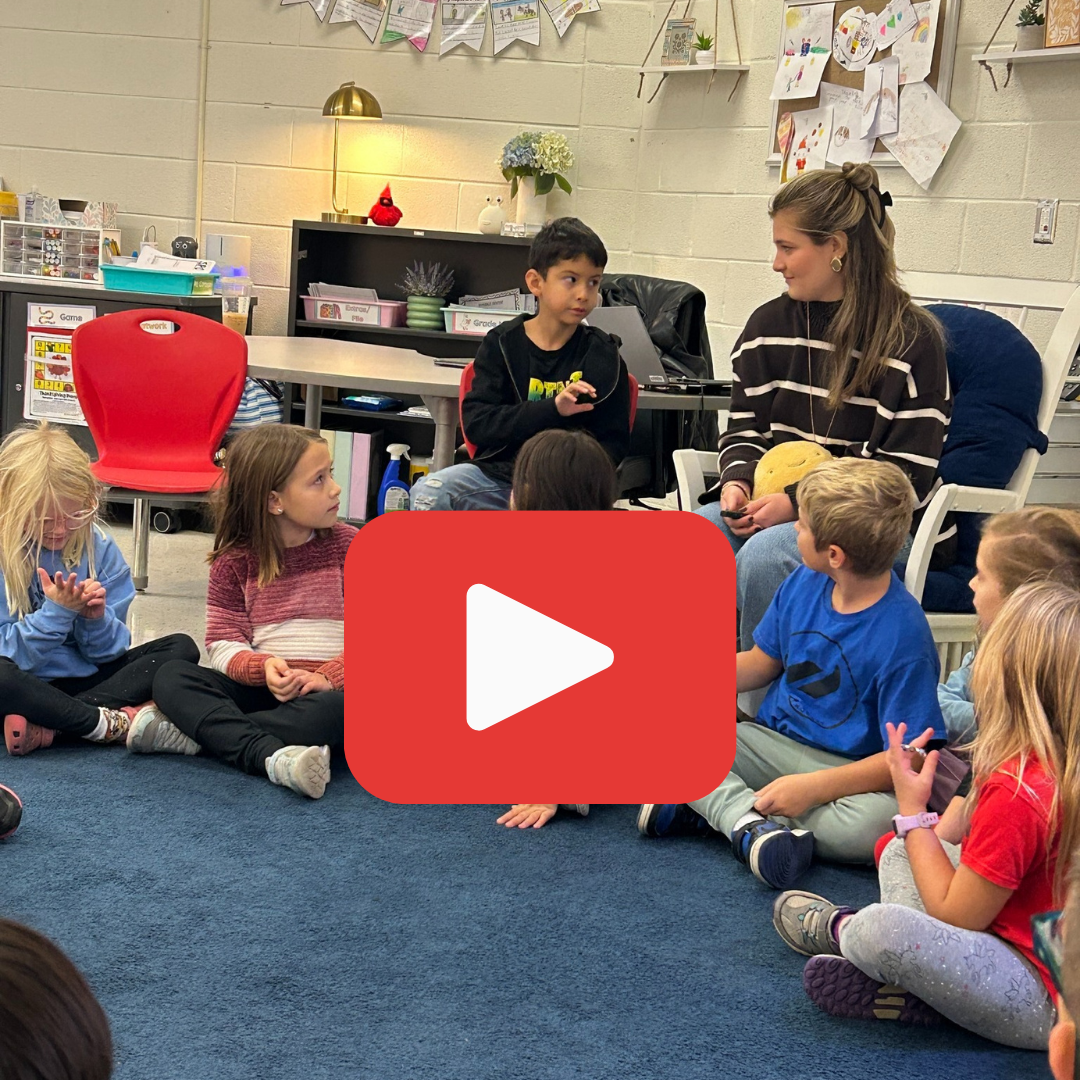 Elementary students and their teacher sit in a circle on a rug for a morning meeting/circle time. A video play button is overlaid.