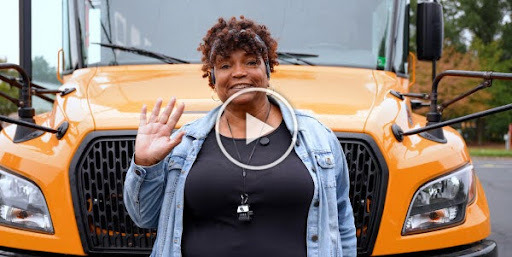 A woman in front of a school bus waves hello