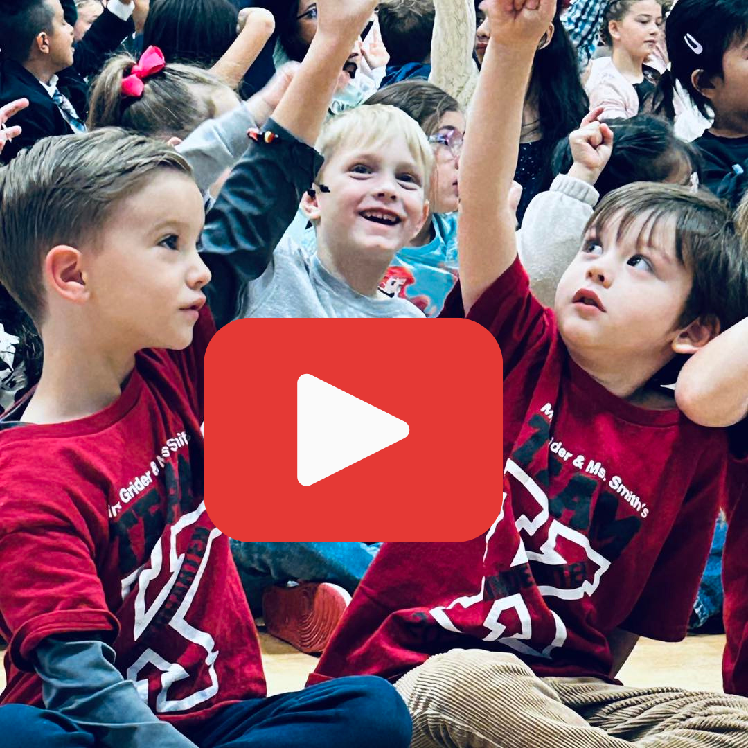 A candid shot of young students in red shirts raising their hands enthusiastically during a school assembly or activity.