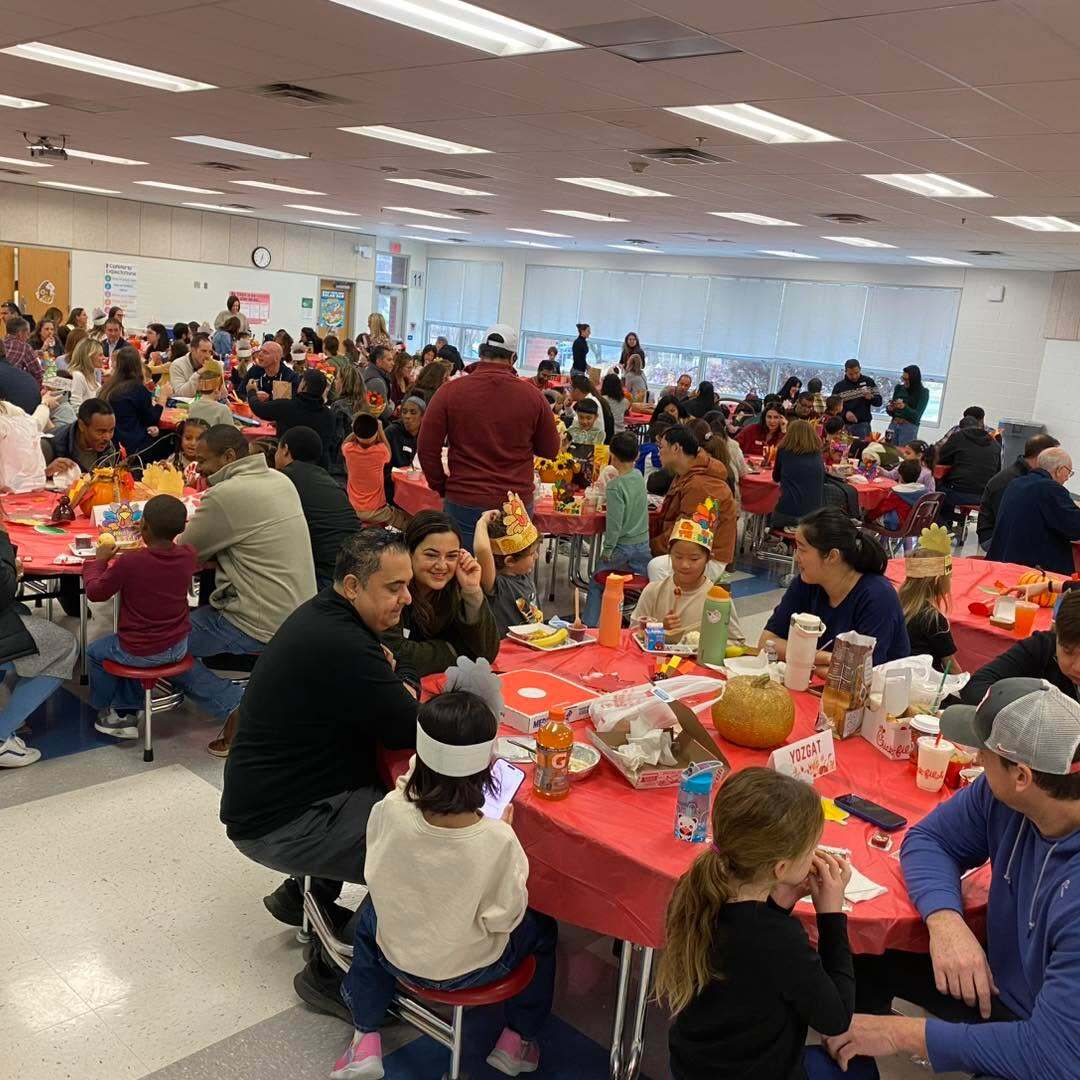 Students and their families are seated in circle tables in a cafeteria enjoying a thanksgiving lunch