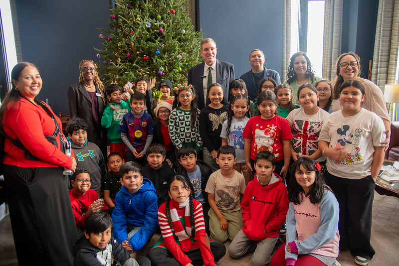 A group of students and teachers pose in front of a festive tree with Senator Warner. 